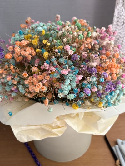 A bouquet of dried baby's breath flowers, dyed in various pastel colors including pink, purple, blue, yellow, and orange, is displayed in a white vase. The flowers are delicate and clustered together, with thin, woody stems. The vase is wrapped in cream-colored tissue paper, which is folded and tucked around the stems. The background is softly blurred, showing a grey wall and a textured white surface on the right. The scene is lit from above, casting subtle shadows. The composition is a close-up shot, focusing on the texture and colors of the flowers. The image does not depict any specific location in Kol'tsovo, Russia.