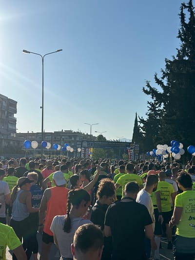 Here's a detailed description of the image:

The image shows a large crowd of people gathered on a city street in Stavroupoli Municipal Unit, Greece. The event appears to be a marathon or similar race, suggested by the banner reading "MARATHONIOS MEGAS ALEXANDROS" (Great Alexander Marathon) and mentions of a 10,000m dynamic walk.  

Many participants are wearing bright yellow or orange t-shirts with the logos "20 YEARS" and "win." Some are also seen in other colored shirts like pink and blue. There is a line of blue and white balloons along the street.

In the background, there are several apartment buildings. Street lights are visible along the street. Above the buildings, a clear blue sky is visible. A large tree is on the left of the image, partially covering the sky.

The crowd is densely packed. The participants are positioned along the race route, ready to start or perhaps already in the midst of the event.