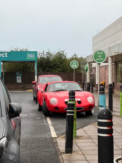 The image captures an outdoor scene in Omagh, United Kingdom, featuring a red sports car parked next to a red SUV in what appears to be a shopping center parking lot. To the left, a grey car is partially visible in the foreground. Overhead, a cloudy sky suggests overcast weather.
Several signs are present in the scene. A turquoise structure on the left displays "Click & Collect" with an accompanying laptop icon. Further to the right, two green circular signs are visible; one reads "Pick up point" and the other displays a taxi icon. Bollards of various colors, including black, blue, and green, are positioned along the pavement.
The ground surface consists of asphalt in the parking area and paved tiles in the pedestrian walkway. The background reveals trees and a modern building with beige cladding. The red sports car, a distinctive classic model, is the primary subject in the foreground, parked adjacent to a newer red SUV.