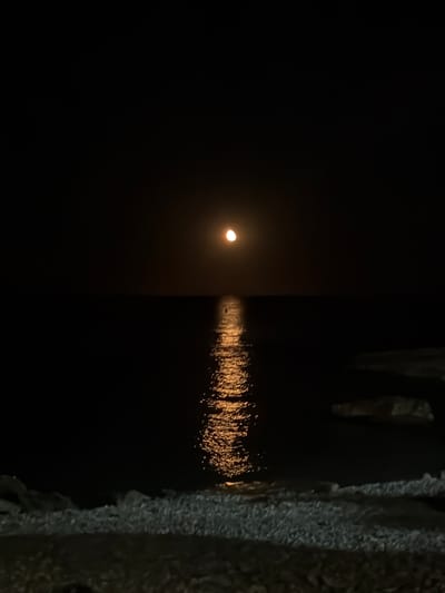 A crescent moon illuminates a dark ocean, casting a shimmering reflection on the water's surface. The shoreline, composed of pebbles and rocks, is visible in the foreground. The image captures a tranquil night scene at a coastal location, possibly within the municipality of Xàbia/Jávea, Spain, given the typical pebbly beaches and clear waters often found in the region. There are no people or identifiable structures present. The overall atmosphere is serene and somewhat mysterious due to the low light conditions.