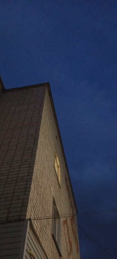 An angled low-light shot focuses on the corner of a light-colored brick building against a dark blue sky. A single rectangular window on the building is illuminated from within, casting a soft yellow glow. Below the brick, a section of the building is clad in horizontal white siding. Faint, reddish graffiti is visible on the brick wall. Thin wires are strung horizontally across the lower right portion of the image. The overall scene suggests dusk or night in an urban setting, possibly Kudymkar, Russia, given the context provided. There are no people or discernable actions taking place.