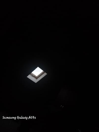 A dark room with a square skylight letting in light from the overcast sky. The skylight is framed with a white trim, and a light brown wooden frame. There is a white "Samsung Galaxy A03s" written in the lower left corner of the image. The location is an unknown room in Hartlepool, United Kingdom. The coordinates given are for the town center of Hartlepool and do not provide information about the specific building depicted.