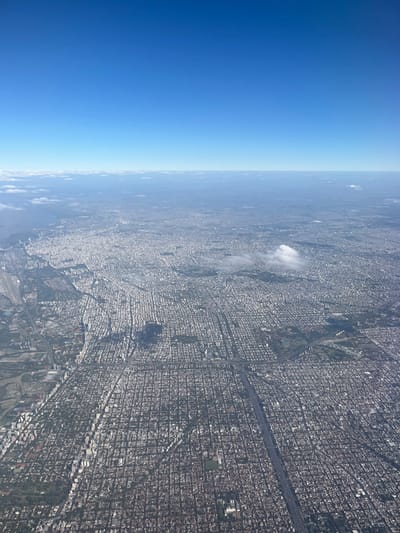 This image presents a high-altitude aerial view of a sprawling urban landscape under a clear blue sky. The scene depicts a significant portion of the northern zone of Greater Buenos Aires, Argentina. The city of Martínez is located within the extensive urban grid visible between the prominent multi-lane Autopista Panamericana (running diagonally from the lower-right to the upper-left of the image) and the Río de la Plata (visible along the far left edge).
The environment consists of a dense, rectilinear grid of urban development, primarily composed of numerous buildings appearing uniformly grey or light-toned from this altitude, indicating residential and commercial structures. Multiple roads and streets dissect this grid, with the Autopista Panamericana being a distinct, wide, multi-lane highway featuring complex interchanges. Interspersed within the urban area are various green spaces, likely parks or recreational fields. The Río de la Plata forms the eastern boundary of the visible landmass, appearing as a vast, calm body of water along the left side of the image. The upper half of the image is dominated by a clear, deep blue sky, transitioning to a lighter blue near the horizon. A few small, scattered cumulus clouds are present over the distant urban area, and a thin line of stratiform clouds marks the distant horizon. The scene is illuminated by bright daylight. No individuals, specific actions, or interactions are discernible due to the elevated perspective.