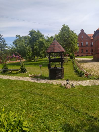 This is a full-shot, eye-level, outdoor photograph taken on a sunny day, featuring a well in the foreground. The well is made of dark wood and has a red, shingled roof. To the left of the well is a paved pathway made of stone. Beyond the pathway, a black metal bench is visible, with a decorative metal archway behind it, holding pink flowers in hanging baskets. A short, dark green bush is planted on either side of the bench. The background reveals lush green grass, trees with full foliage, and a large, red brick building with a tiled roof and multiple windows. The building appears to be historic, with a pitched roof and architectural details typical of older European structures. The sky is partly cloudy, with wisps of white clouds against a light blue background. The scene appears to be a well-maintained garden or courtyard area, possibly part of a historical site or estate. The location is identified as the Gusevsky Museum of Local Lore in Gusev, Russia.