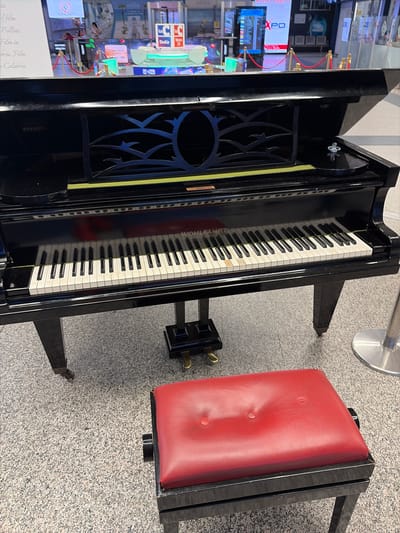 The image shows an upright piano and a red piano bench. The piano is black and appears to be made of wood. It has a keyboard with black and white keys, and a decorative grill on its left side. The bench is upholstered in red leather and has a black metal frame. The piano and bench are placed on a speckled grey floor. In the background, on the left side of the image, there are several screens displaying various images and text, suggesting a public space or exhibition. One screen shows a video with text that appears to be in Italian, including "POLLINI" and "FILM IN CALABRIA". The lighting suggests it is daytime, and the setting could be indoors in a museum, gallery, or public hall in Lamezia Terme, Italy. There are no people visible in the image. The overall mood is one of stillness and exhibition.