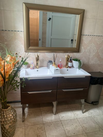 The image shows a bathroom sink with two basins and a brown wooden cabinet with chrome accents. The sink is located in Buea, Cameroon at the coordinates (latitude: 4.145, longitude: 9.23). The basin is filled with assorted toiletries including a bottle of hand soap and a pump bottle of lotion. The cabinet has two drawers, both of which are closed. The sink is situated in front of a large, framed mirror with a gold trim. Above the sink is a light beige tile wall with an elaborate, repeating, floral pattern. To the left of the sink is a vase filled with artificial orange flowers. The floors are tiled in a light beige color. In the right corner is a black trash can with a gray liner. The bathroom appears to be relatively clean. The background of the image is a light-colored wall with a doorway visible in the reflection of the mirror.