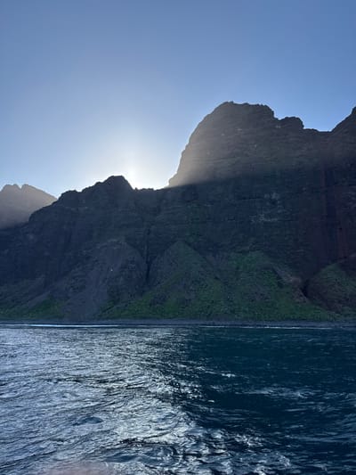 The image displays a rugged mountainous coastline viewed from the ocean near the city of Unknown, United States of America. The upper portion of the frame features a clear, light blue sky. Below this, a range of dark, imposing cliffs and peaks extends across the horizon, appearing silhouetted. The sun is partially obscured by one of the central mountain peaks, generating a powerful backlight and visible rays of light extending downwards and outward from behind the peak. The mountainous terrain exhibits steep, shadowed rock faces with some visible green vegetation on the lower slopes, particularly along the water's edge. The lower half of the image is dominated by dark blue ocean water with a textured, choppy surface, reflecting significant sunlight in bright streaks on the left. The water meets the base of the cliffs along a narrow, dark strip of shoreline. No people, animals, or man-made objects are visible.