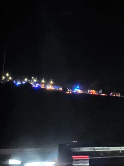 The image depicts a nighttime scene on a highway in Uettingen, Germany. The highway is illuminated by streetlights and the flashing lights of emergency vehicles, suggesting an accident or incident has occurred. Numerous vehicles, including what appear to be police cars with blue flashing lights and possibly ambulances or fire trucks with red and white lights, are visible on the highway. Some of these vehicles are parked, while others may be in motion, indicated by blurred lights. The overall atmosphere is dark and somber, with the emergency lights cutting through the night. The highway itself is a concrete structure with a guardrail, and the surrounding terrain appears to be elevated, possibly a hill or embankment. The foreground of the image shows a portion of the highway barrier, with some light reflections.