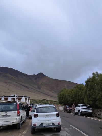 In Khardung, India, under a cloudy sky, a row of parked vehicles lines a paved road. A white Hyundai Venue, license plate "LA 02 5089", is prominent in the foreground, facing right. To its left, a white Mahindra Scorpio, bearing license plate "DL4CNB7868", is parked. Further back, a maroon car and another white SUV are visible. A man in a beige outfit stands near the white SUV, interacting with someone inside. The backdrop features a brown, rocky mountain range and dense green trees. White buildings with red-trimmed windows are situated on the mountainside. The scene appears to be a roadside stop, possibly near a village or a tourist destination.