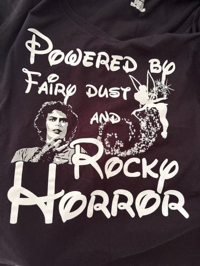 A black t-shirt with white text and graphics is laid flat. The t-shirt features the text "POWERED BY FAIRY DUST AND ROCKY HORROR". To the left of the text is a grayscale image of Tim Curry as Dr. Frank-N-Furter from "The Rocky Horror Picture Show". To the right of the text is a silhouette of Tinkerbell, a fairy from Peter Pan, sprinkling stardust. The t-shirt is not in the city of Červená Píska, Czechia; it is a piece of clothing.