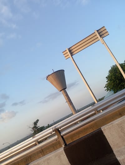 This is a low-angle, wide shot of a water tower and a sign structure in Gungi, Nigeria. The sky is a clear blue with scattered white clouds, indicating it is daytime. The sun is low in the sky, casting warm light on the scene, suggesting it is either early morning or late afternoon.

The main subjects are the conical water tower, supported by a cylindrical concrete column, and a large, angled sign structure composed of horizontal metal slats supported by two diagonal beams. Both structures are weathered and appear to be made of concrete and metal.

In the foreground, there is a concrete barrier with metal railings, typical of a roadside or bridge. The railings are made of several parallel silver pipes. The concrete barrier is rough and shows signs of wear.

In the background, there is a treeline with lush green trees, and further in the distance, a hazy landscape with some visible structures and a street light. The overall impression is one of an outdoor, possibly urban or semi-urban, environment. There is no text visible in the image. The lighting and composition create a slightly dramatic and perhaps solitary mood.