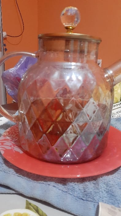 An indoor, close-up image focuses on a clear glass teapot with a faceted diamond pattern on its body, C-shaped handle, and spout. The teapot contains a reddish-brown liquid. Its clear lid features a gold-colored band and a faceted clear glass knob. The teapot rests centered on a solid red-orange ceramic plate adorned with a partial white floral design along its rim. This plate is positioned on a light blue terry cloth towel. The background consists of a plain orange wall, with a white electrical outlet and black wires visible on the upper left, and a blurred purple plastic bag behind the teapot's handle. To the right, another blurred, light-colored object and patterned fabric are partially visible. In the immediate foreground, a portion of a white plate with painted yellow lemon slices and green leaves is seen, alongside a sliver of white packaging. No people are present. Specific identifying features for the city of Pap, Uzbekistan, are not discernible within the image.