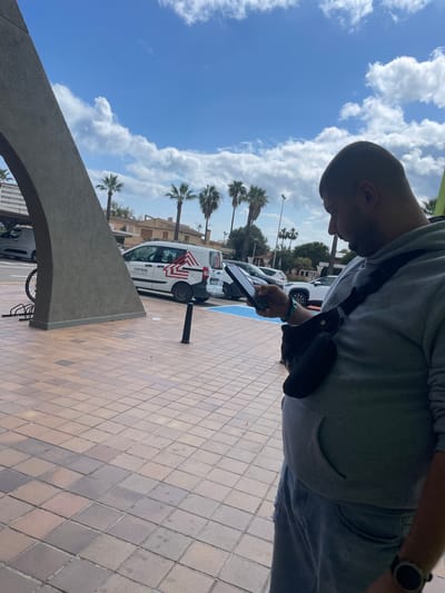 An outdoor scene in Sant Llorenç des Cardassar, Spain, is depicted under a bright blue sky with scattered white cumulus clouds. In the immediate foreground, a male subject is visible from the waist up, positioned in profile facing right, actively viewing a black smartphone held in both hands. He is dressed in a grey hooded sweatshirt, light blue distressed jeans, a black cross-body bag slung across his chest, a black smartwatch on his left wrist, and a blue/green beaded bracelet on his right wrist.
To the left, a large, grey, textured concrete architectural element, forming a partial arch, dominates the left portion of the image. Behind this structure, a black metal bicycle rack is partially visible. The ground is paved with square tiles in varying shades of brown and grey. A small black bollard and a piece of white debris are present on the tiled pavement in the midground.
Beyond the pavement, a street is visible with several parked vehicles. A white commercial van, bearing the "coemsa" logo and a red and white striped graphic, is parked in a blue-marked parking space. Other vehicles, including a white SUV and a dark car, are partially visible. The background features multiple palm trees interspersed with low-rise buildings characterized by light-colored facades and tiled roofs. A streetlamp is also visible among the trees.