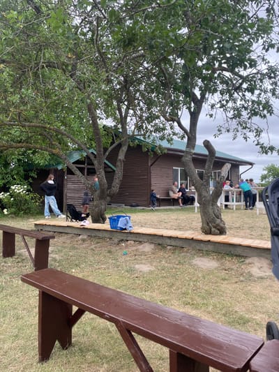 The image depicts an outdoor scene at a rural establishment, likely a cafe or guesthouse, in or near the Ратайчыцкі сельскі Савет, Belarus. In the foreground, two brown wooden benches are visible, one in sharp focus and the other further back. Behind the benches, a wooden walkway with a light-colored plank surface runs horizontally across the image. Beyond the walkway is a grassy area with scattered patches of dry soil.

A two-story wooden building with a green corrugated metal roof serves as the backdrop. The building has windows and a wooden facade. Several people are gathered in front of and on the porch of the building. On the left side of the image, near the building, a young woman in a black hoodie and light blue jeans stands next to a black stroller. Further to the right, several individuals are seated on benches and at tables on the porch. A child in a blue shirt is standing on the grass to the left of the seated group. On the far right, a group of people are standing near a white railing.

Several trees with green foliage are scattered throughout the scene, with a particularly large tree in the center foreground. In front of the building, on the wooden walkway, a blue bucket with a light blue cloth draped over it is visible. The sky is overcast with a cloudy, grayish hue. The overall impression is of a casual, outdoor gathering space.
