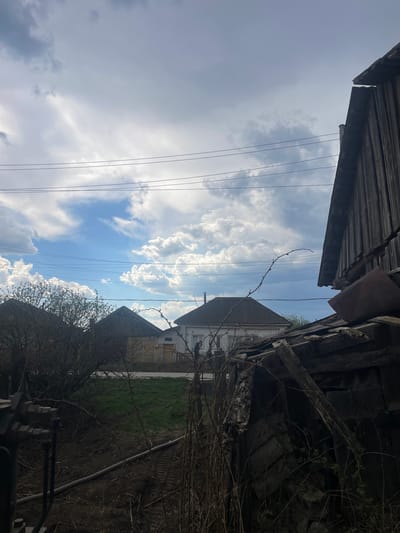 The image, taken from a low angle in Поедуги, Russia, shows a view upward towards a partly cloudy sky. Power lines stretch across the sky. Overhanging wooden structures, possibly parts of buildings or sheds, frame the top and right sides of the image. Two houses are visible in the middle ground, separated by a patch of green grass. In the foreground, bare tree branches and some obscured objects add depth to the scene. The overall impression is one of a rural or semi-rural environment with a mix of natural and man-made elements.