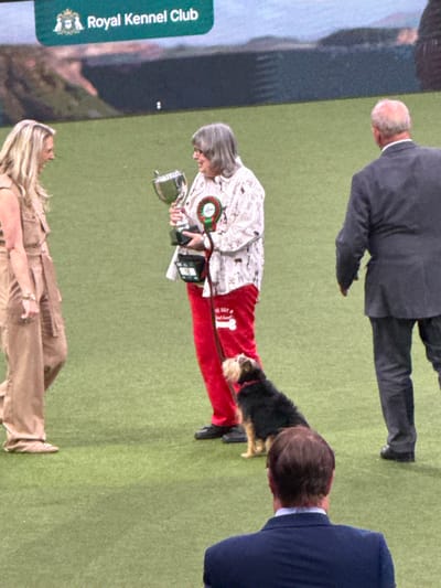 This image depicts an indoor award ceremony at the National Exhibition Centre (NEC) Arena in Solihull, Metropolitan Borough of Solihull, United Kingdom, identified by the "Royal Kennel Club" banner displayed on a large screen in the background.
Four individuals and one dog are present on a green carpeted area. On the far left, a woman with long blonde hair, wearing a tan sleeveless jumpsuit, stands smiling and facing the center. In the center, an older woman with grey hair and glasses holds a silver trophy cup in her left hand and a smaller dark trophy base in her right. She wears a white long-sleeved shirt patterned with dog silhouettes, red trousers with a "WE GOT" and bone graphic on the left thigh, and black shoes. A red and green rosette is pinned to her chest, and she holds a red leash that extends to a black and tan terrier-type dog sitting patiently beside her. The dog wears a pink collar and looks up and slightly left. To the right of the central woman, a bald man in a dark grey suit walks away, his back partially turned to the camera. In the foreground, with his back to the viewer, a man with brown hair wears a dark blue suit jacket, observing the scene. The background features a large screen displaying the "Royal Kennel Club" logo and a blurred landscape image beneath it. The floor is a uniform green carpet.