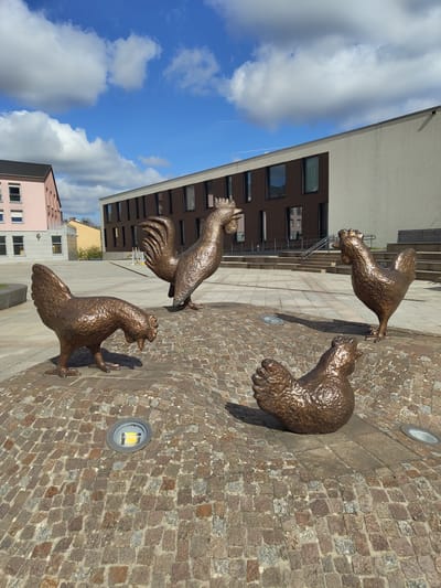 The scene depicts an outdoor public plaza in Bertrange, Luxembourg, under a bright blue sky with scattered white cumulus clouds. Four bronze statues of chickens and roosters are positioned on a multi-textured paved surface. The foreground and central area feature irregular reddish-brown cobblestones, while larger, rectangular grey flagstones define walkways and elevated sections.

One hen statue in the immediate foreground, positioned left of center, faces right with its head lowered as if pecking the ground. Another hen statue is situated in the mid-ground, right of center, facing left. A large rooster statue stands prominently in the mid-ground, towards the rear-left, facing left. A fourth hen or smaller rooster statue is visible in the background, towards the far-right, facing left. Two circular ground lights are embedded in the cobblestones, one emitting a yellow light near the hen statue in the foreground.

In the background, two multi-story buildings are visible. To the left, a light pink building with a dark roof and white window frames rises. To the right, a larger, modern building with a light grey facade and contrasting dark brown vertical panels houses multiple rectangular windows. A wide set of grey steps with a metal handrail leads up to an elevated section of this building, where a wooden bench is integrated. Shadows from the statues and buildings indicate direct sunlight from an overhead source. No people are present in the image.