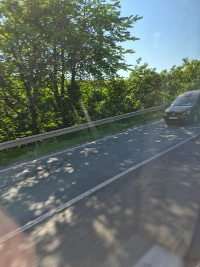 The image captures a roadside scene with lush green trees lining a curved road under a clear blue sky. A dark-colored van is visible driving on the right side of the road, its front bumper and headlights illuminated by sunlight. The road itself is asphalt with a white line marking its edge. Shadows of tree leaves are dappled across the road, indicating a sunny day. A metal guardrail runs along the left side of the road, separating it from the vegetated embankment. The overall impression is of a scenic route, possibly on the outskirts of a town or city. Given the information that this location is in Fălticeni, Romania, this scene likely depicts a road within or near that city, surrounded by its natural environment.