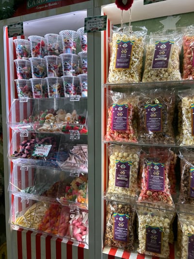 This image captures a vibrant display of sweets and flavored popcorn, likely within a candy store or market stall in Chalkida, Greece. The scene is presented with a slight rotation to the left.
The upper shelf showcases numerous transparent bags of gourmet popcorn, neatly arranged in rows. Visible flavors include "Extra Butter," "Chocolate Flavor," and "Strawberry Cream." Each popcorn bag features a label with a colorful logo incorporating a jester's hat and the text "Cirque du Bubble."
Below, a lower shelf features an array of bulk candies displayed in clear, gravity-fed dispensers. These dispensers hold various confections, such as colorful gummy candies, marshmallow-like sweets, and possibly liquorice or other chewy candies. To the left of these dispensers, clear plastic cups are stacked, each containing a small packet of candy and bearing the brand name "Bubble Tale."
Notable textual details include a green sign on the left with Greek text "ZAXAPΩTA XWPA" (likely "Sweets Country" or similar) and pricing information "2,20€ / 100gr." Another green sign below it clearly states "Candy" and "Cirque du Bubble." The backdrop features a light wall with a subtle star pattern on the left and cheerful red and white vertical stripes on the right, suggesting a playful, carnival-like atmosphere. No people are visible, and the time of day or weather conditions cannot be determined.