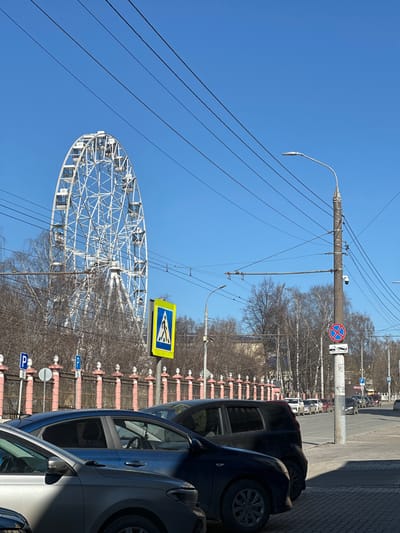 The image shows a street scene in Postol, Russia, featuring a Ferris wheel. The Ferris wheel is white and located on the left side of the image, with a clear blue sky in the background. Overhead power lines stretch across the top left of the image. The right side of the image shows several parked cars along a street with a sidewalk. A pink and white fence separates the sidewalk from a line of trees. Street signs are visible, including a "No Stopping" sign and a "Pedestrian Crossing" sign. Parking signs are also located behind the cars. The image is taken during the daytime with clear sunlight.