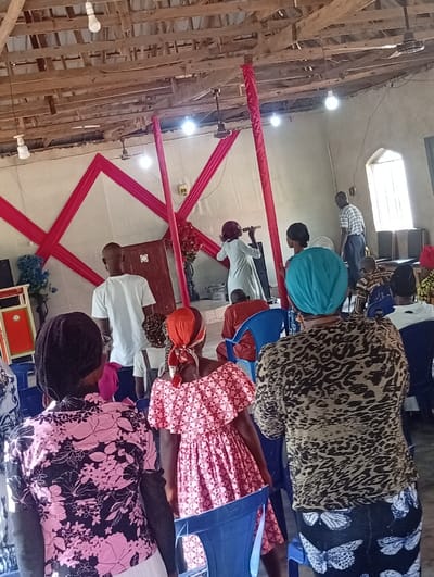The image displays an indoor communal gathering in Isinyi-Nando, Nigeria. The environment features a raw, exposed wooden beam and plank ceiling with multiple bare light bulbs and two ceiling fans. Light-colored walls are visible, with the back wall adorned by red fabric drapes arranged in a geometric criss-cross pattern. Similar red fabric is wrapped around two vertical supports in the central area. A tall, arched window is present on the right wall.
In the foreground, viewed from behind, are three individuals: on the left, a person wearing a black top with pink floral patterns; in the center, a person in a pink and white patterned off-the-shoulder dress with a red headscarf, bending towards a blue plastic chair; and on the right, a person wearing a brown and black leopard-print top and a dark skirt with white butterfly patterns, topped with a teal headscarf.
In the midground, a woman in a light-colored, long-sleeved garment holds a microphone in her right hand, oriented towards the right side of the frame, positioned in front of a dark wooden structure that appears to be a lectern. To her left, partially obscured, a man in a white t-shirt stands with his back mostly to the viewer. Another individual, wearing an orange top, is partially visible kneeling or bent over near the center. Black audio speakers are positioned on the left side of the scene.
In the background, near the arched window, a man in a light-colored striped shirt and dark trousers stands. Numerous blue plastic chairs are scattered throughout the room, some occupied by seated individuals further back, and some unoccupied in the foreground. A bouquet of red flowers is visible near one of the red-draped vertical supports.