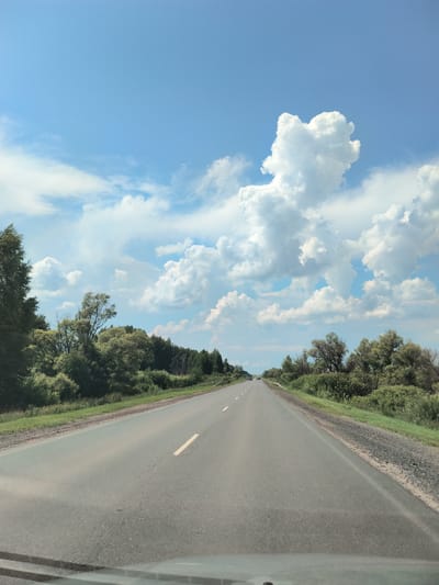 The image depicts a road passing through a rural landscape under a cloudy sky. The road is asphalt with a yellow line marking the lane. Dense green trees and vegetation line both sides of the road. In the distance, a few cars are visible. The sky is a vibrant blue with large, fluffy white clouds. The scene suggests a sunny day, possibly in the summer, with the road appearing to lead towards the horizon. Based on the context, the location is identified as Старокутлумбетьево, Russia.