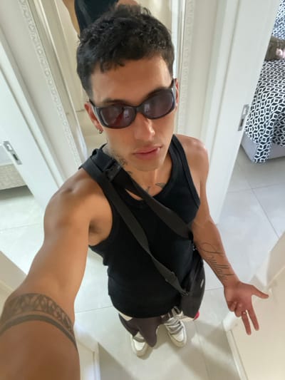 An individual, identified as male with a dark skin tone, dark curly hair, and wearing dark sunglasses, is depicted indoors in what appears to be a residential or hotel setting, reportedly located in Bombinhas, Brazil. The subject is wearing a black tank top, dark pants, and light-colored sneakers. A black shoulder bag with a visible plastic buckle strap crosses the chest from the left shoulder. Tattoos are present on the left forearm (a wide, dark tribal band), on the right forearm (script lettering, partially legible as "Trullaby"), and possibly on the neck/collarbone area. A silver-colored earring is visible in the left earlobe, and a ring is present on the left hand. The individual is positioned centrally, taking a high-angle selfie, with the left arm extended towards the camera. The right hand, lowered to waist level, displays a "peace sign" or "V" gesture with the index and middle fingers extended.
The immediate environment consists of white walls and light-colored tiled flooring. To the right, an open doorway reveals a partial view of an adjacent room containing a bed with a black and white geometric patterned duvet or bedspread. To the left, a tall, white, decorative doorframe or mirror frame is visible, reflecting a distorted image of the subject's upper body. The lighting is consistent with an indoor setting.