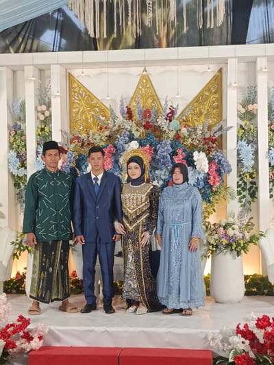 Four individuals are formally posed on a decorated stage, facing the camera at an event in the city of Kedung Dowo, Indonesia.
From left to right:
1. A man with dark skin, wearing a black peci (cap), a dark green long-sleeved shirt with white geometric patterns, a patterned sarong over light trousers, and light brown open-toed sandals.
2. A younger man, wearing a dark blue suit with a white shirt and blue tie, and black dress shoes. His left arm is positioned close to the woman beside him.
3. A woman, wearing an elaborate dark blue or black traditional gown extensively decorated with gold and silver embroidery and fringe, a dark head covering (hijab), and a prominent ornate gold headpiece. She has visible makeup and wears silver open-toed shoes. Her left hand is near the man's arm.
4. A woman, wearing a light blue long-sleeved dress with a belted waist, a dark head covering (hijab), gold bangles on her right wrist, and dark open-toed sandals.
The individuals stand on a raised white-covered stage. The backdrop consists of a white wall adorned with large gold, crown-like ornamental panels in the upper center, flanked by voluminous arrangements of artificial flowers in shades of blue, pink, red, and white. Smaller white pendant lights hang from the top of the backdrop. On the stage floor, large white vases containing floral arrangements are positioned to the sides. Above the stage backdrop, a dark reflective surface is visible, from which white decorative strands hang. A light blue fabric canopy is partially visible at the top left. A red low wall or step is present at the very bottom front of the stage.