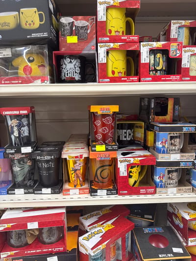 The image shows a retail shelf displaying a variety of merchandise, predominantly mugs and glasses, featuring popular anime and manga franchises such as Pokémon, Naruto, One Piece, and Death Note. The items are neatly arranged in their original packaging. The lighting suggests an indoor retail environment, likely a store. The price tags visible indicate prices in Euros, with some examples including "12,99€", "9,99€", and "19,99€". The merchandise is predominantly displayed on two shelves, with a variety of colored boxes and packaging. The overall scene is a typical retail display of licensed merchandise.