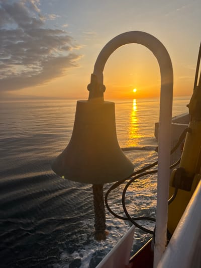 This image captures a serene sunset over a calm ocean, viewed from the deck of a ship. In the foreground, a ship's bell, made of dark metal, hangs from a curved support. The bell has a thick rope attached to it, used for striking. To the right of the bell, a yellow metal pole with some wiring is visible, part of the ship's structure. The sun, a bright orange orb, is setting on the horizon, casting a shimmering golden path across the water. Wispy clouds, tinged with orange and yellow from the sunset, dot the sky. The ocean surface is rippled, with white foam visible in the foreground, indicating the ship's movement. The overall scene is one of tranquility and the vastness of the sea at dusk. The location is not specified in the image, and no people are visible.