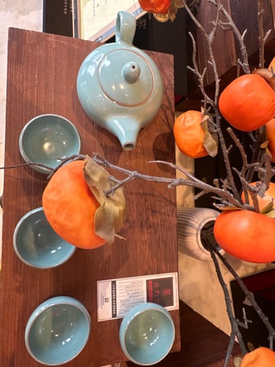 The image captures a static indoor display featuring artificial persimmons, ceramic tea ware, and wooden elements. In the foreground, a light blue-green glazed ceramic teapot, characterized by its round body, short spout, and handle, rests on a dark reddish-brown wooden plank. Five small, matching blue-green ceramic cups are arranged around the teapot on the same wooden surface.

Above the tea set, bare, gnarled branches are adorned with vibrant orange artificial persimmons, many still bearing their green or brown calyxes, evoking an autumnal or harvest theme. These branches appear to originate from vases, with a light-colored, fluted vase partially visible holding some branches in the background. Further back, a darker wooden surface and a black box or packaging are discernible.

A white rectangular label or card, placed on the wooden plank near the cups, features Chinese characters and some English text. Visible text includes "JUN PORCELAIN", "盧鈞家" (Lu Jun Jia), and "窑" (kiln). Key phrases like "140年道統傳承-钧窑復興鼻祖" (140 years of traditional inheritance - the ancestor of Jun porcelain revival) are present, along with "年3月18日" (March 18th) and "当百年世家" (When a century-old family/clan). This text strongly suggests the display is related to the heritage and craftsmanship of Jun porcelain, a type of Chinese ceramic ware, likely situated in a shop, showroom, or cultural exhibit in Fangshan District, China. The color palette combines the bright orange of the fruit with the serene blue-green of the ceramics and the warm browns of the wood, creating an aesthetically pleasing arrangement.