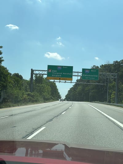 The image depicts a highway scene in Glen Burnie, United States of America, with overhead signage directing traffic. On the left, a green sign indicates "195 EAST BWI Thurgood Marshall Airport" with an "EXIT 12 ONLY" designation below. To the right, another green sign reads "195 WEST Catonsville" and states "EXIT 1/2 MILE". The road is a multi-lane highway with white lane markings. Trees and foliage line the sides of the highway. The sky is clear blue with a few scattered white clouds. The reflection of a red vehicle is visible in the foreground.