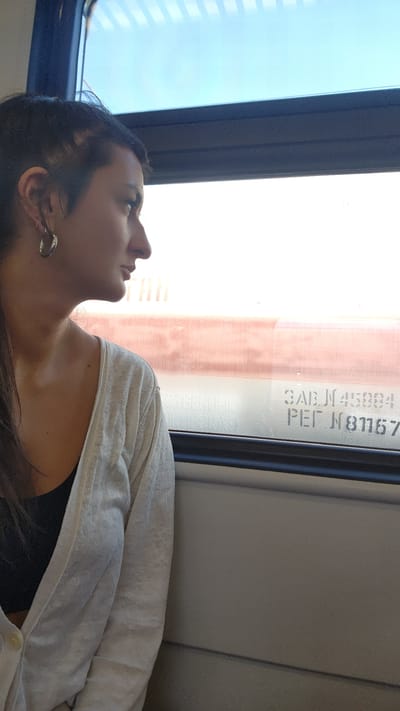 A woman with dark hair is looking out of a train window. She is wearing a black tank top and a light-colored cardigan. Outside the window, a train car with Cyrillic lettering reading "SABJ N45994 PEI N81167" is visible. The background shows a blue sky with some clouds. There is no information present to indicate this image was taken in Lukhovitsy, Russia.