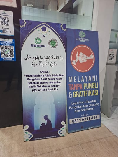 The image shows a street view in Lampeuneurutblang, Indonesia, with two large blue banners placed vertically on the ground against a wall. The banner on the left features the RSUD Meuraxa logo and a quote in Arabic script, followed by its Indonesian translation and a reference to the Quran. The banner on the right, also branded with RSUD Meuraxa, prominently displays the message "MELAYANI TANPA PUNGGLI & GRATIFIKASI," which translates to "SERVING WITHOUT BRIBERY & GRATIFICATION." It also includes instructions to report any illegal collections or gratuities and provides a contact number, 0811 6711 414. Below the contact number, a website address and social media icons are visible, along with the handle "rsmeuraxa." Both banners are decorated with Islamic-inspired patterns. To the left of the banners, there are two smaller blue flyers with QR codes, one for customer feedback and another for health information, both also referencing RSUD Meuraxa. A small white box with a QR code and text "MELAYANI PESERTA ASURANSI PLN" (serving PLN insurance participants) is also visible to the right of the main banners. The ground is tiled with light-colored square tiles.