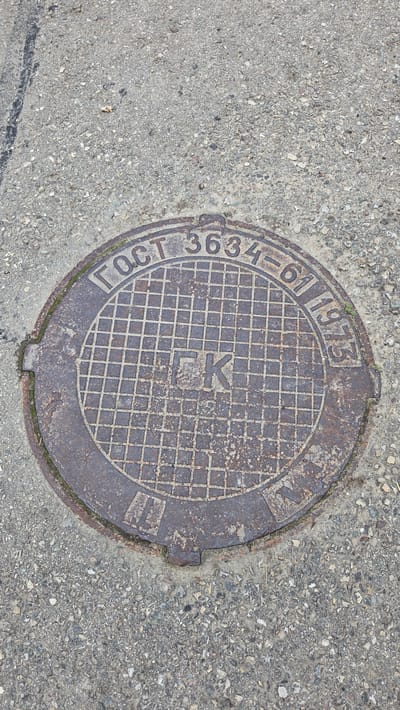 The image shows a close-up, overhead view of a cast iron manhole cover set into a concrete pavement. The manhole cover is circular and has a textured, grid-like pattern across its surface. In the center of the grid, the Cyrillic letters "LK" are embossed. Around the perimeter of the manhole cover, text is inscribed. The visible text reads "ГОСТ 3634-61 1973". This inscription indicates that the manhole cover conforms to the GOST 3634-61 standard, and was manufactured in 1973. The manhole cover is weathered and shows signs of rust and wear. The surrounding concrete is rough and speckled with small stones and debris. The lighting suggests it is daytime, with no specific weather conditions evident. The location is identified as Ūlupji, Latvia. The image focuses solely on the manhole cover and its immediate surroundings, creating a utilitarian and somewhat stark aesthetic.