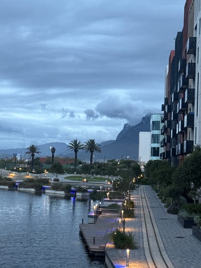 The image shows a waterfront scene, potentially a river or canal, bordered by a paved walkway with benches and palm trees. On the opposite side of the water, there is a row of buildings. One building is a modern structure with a checkered façade, and another is a white building. The sky is cloudy and overcast, and a mountain range is visible in the background, partially obscured by clouds. The location could be in Milnerton, South Africa.