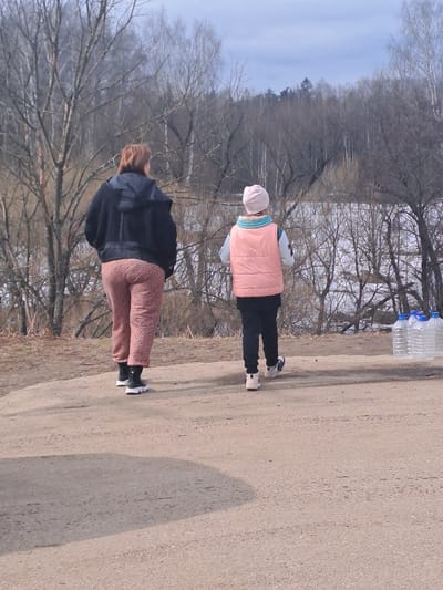 The image displays an outdoor scene in Avdot’ino, Russia, under daylight conditions, likely in late winter or early spring due to the bare trees. A dirt or gravel path extends from the foreground into the midground. Two individuals are walking away from the camera on this path.

The person on the left, an adult, has reddish-brown hair and is wearing a black hooded jacket, pinkish-brown patterned pants, and black and white athletic shoes. The person on the right, appearing to be a child or smaller adult, wears a light pink beanie, a light blue neck gaiter, a light pink sleeveless puffer vest over a light-colored long-sleeved shirt, black pants, and light-colored athletic shoes. Both individuals are moving in the same direction, away from the viewer.

To the right of the path, positioned near a line of bare trees, are four transparent 5-liter plastic bottles with blue caps, standing upright and filled with a clear liquid. Beyond the immediate treeline of bare deciduous trees, a body of water is visible, bordered by a denser treeline in the distance, all under a pale, cloudy sky.