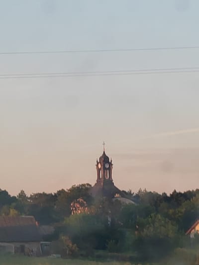 The image depicts a landscape view of the village of Volchinsk, Volchinski Selsoviet, Kamenets District, Brest Region, Belarus. In the center of the frame, a church tower with a clock and a cross stands tall above a dense canopy of green trees. To the left of the church, a portion of a building with a dark, sloped roof is visible, and to the right, another building with an orange roof is partially seen. The sky above is a soft gradient of pale blue and peach, suggesting either sunrise or sunset. Two thin, horizontal lines, likely power lines, traverse the upper portion of the sky. The overall impression is one of a serene rural setting dominated by the prominent church structure.
