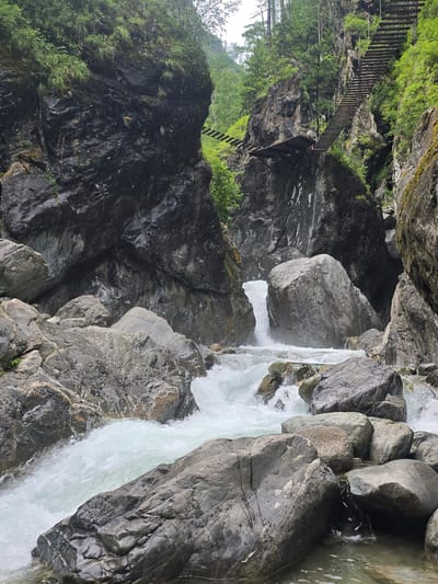 The image depicts a rugged natural landscape featuring a turbulent river flowing through a narrow gorge. Large, dark, wet rocks dominate the foreground and midground, with white water crashing and churning over them. The gorge walls are steep and rocky, covered in patches of green vegetation and moss. In the background, a wooden suspension bridge is visible crossing the gorge, and a set of wooden stairs ascends a steep, rocky cliff face. The overall environment suggests a wild, natural setting with evidence of human infrastructure for access. The location is identified as Arshan, Russia. There are no people visible in the image.
