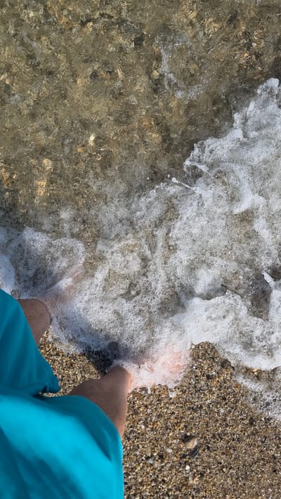An overhead photograph depicts a person's lower legs and feet partially submerged in the shallow, clear waters of a beach in Asprovalta, Greece. The individual is wearing turquoise swim shorts, visible on the lower-left quadrant of the frame. A white, foamy wave breaks around their feet, creating numerous bubbles and splashes. The seabed, composed of small, varied-colored pebbles and coarse sand, is clearly visible through the transparent water, particularly in the upper portion where the water is less agitated. The shoreline transitions from wet pebbles to drier ones further up the beach. The scene is brightly illuminated by natural daylight.
