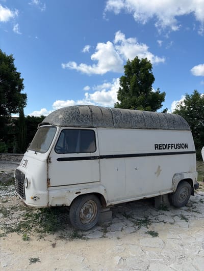 A white, vintage van with the word "REDIFFUSION" in black lettering on its side is parked outdoors. The van is old and appears weathered, with rust visible on some parts. It is situated on a ground covered with sand and stone, with grass growing in patches. In the background, there are trees and a blue sky with white clouds. The image seems to be taken during the daytime. No people are present in the image. The location is identified as Semogorskiy, Russia.