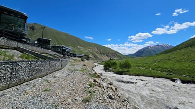 This image captures a scenic mountain landscape under a bright blue sky with scattered white clouds. In the foreground, a rocky, gravelly embankment lines a fast-flowing, muddy river. To the left, a modern, dark-colored building complex with large glass windows is built into the hillside, supported by gabion walls filled with stones. Several dark, multi-story structures, possibly hotel rooms or cabins, are visible along the terraced slope. The building's design suggests a contemporary architectural style, contrasting with the rugged natural environment.
Beyond the structures, a steep, grassy mountain slope dominates the left side of the frame, showing signs of erosion in places. A road or path is visible winding up the mountain. The river flows from the mid-ground towards the right, where it enters a wider valley. The right side of the valley is characterized by lush green meadows dotted with rocks and a few small trees. In the distance, a snow-capped mountain peak stands majestically against the clear sky, with some power lines visible on the lower slopes of the mountains.
The time of day appears to be midday, given the strong sunlight and the bright illumination of the landscape. The weather is clear and sunny, with puffy clouds suggesting a pleasant, temperate climate. The location cues, such as the mountainous terrain, the type of vegetation, and the architecture, are consistent with a remote, possibly tourist-oriented, area in a mountainous region like Kamskho, Russia. The overall mood conveyed is one of natural beauty and modern infrastructure coexisting in a dramatic mountain setting. No visible text is present in the image.