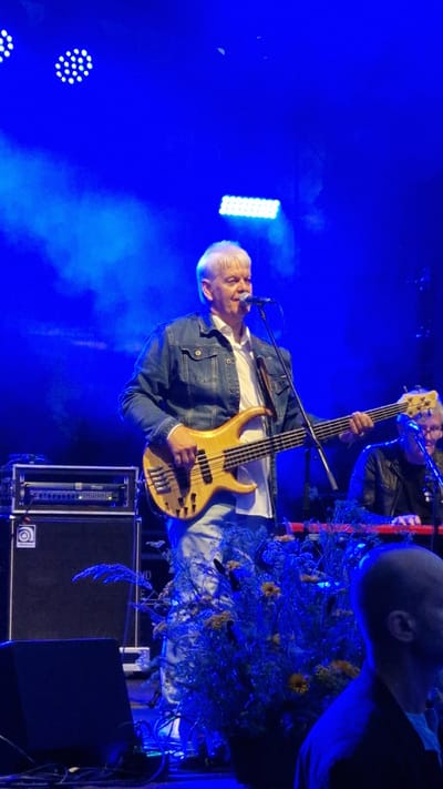 A middle-aged white male with short, grey hair is on a stage under blue lighting, playing a light-colored bass guitar and singing into a microphone. He is wearing a white t-shirt and a denim jacket. To his right, another person wearing glasses and a dark jacket is playing a keyboard. The stage is decorated with a large arrangement of blue and yellow flowers. In the background, there are stage lights and sound equipment. The image appears to be taken in Priekule, Latvia.