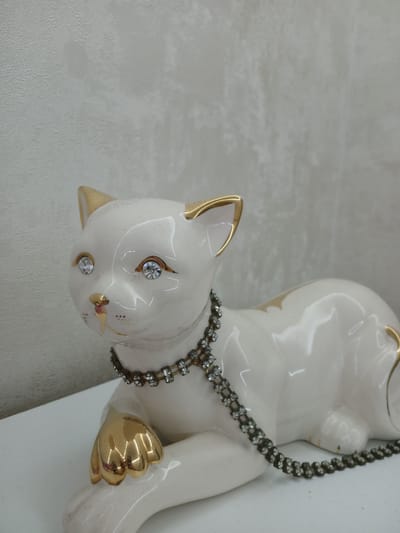 A white ceramic cat figurine with gold accents and rhinestone eyes and collar is lying on a white surface against a textured grey wall. The figurine is positioned on the left side of the frame, with its head turned to the left. The cat's ears, nose, and paws are accented with gold. A two-strand rhinestone collar encircles the cat's neck. The background is a plain, textured grey wall. The scene appears to be an indoor setting. There is no indication of the location being in Sabail Raion, Azerbaijan.
