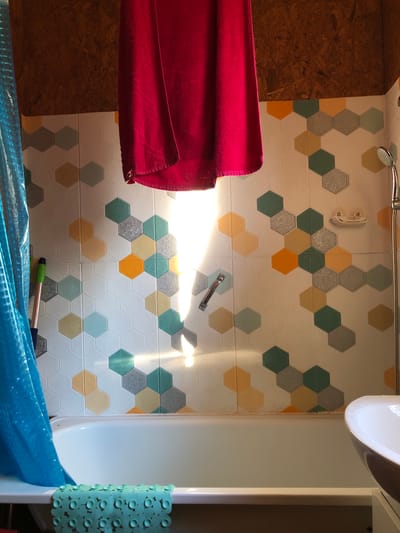 In a bathroom in Izdeshkovo, Russia, a red towel hangs over a bathtub. The walls are tiled with a honeycomb pattern in white, yellow, gray, and teal. A blue shower curtain is to the left of the bathtub. A mint green bath mat with suction cups sits on the edge of the tub. A shower head is visible on the right side of the wall. The scene is lit by sunlight streaming through a window, creating bright reflections on the wall and tiles. The ceiling is made of exposed particle board.