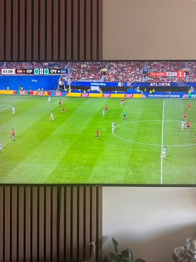This image shows a television screen displaying a soccer match. The score is 0-0 between ESP and CPV. The time on the clock is 61:09. The match appears to be taking place in Atlanta, as indicated by the text on the screen. The game is being broadcast live. In the background of the television screen, a stadium filled with spectators wearing red and white clothing is visible. The foreground of the image shows the green soccer field with players in white and red uniforms. There are also advertising boards around the field, featuring the Lay's logo. The television itself is mounted on a wall, with a decorative wooden paneling to the left and a plain white wall to the right. In the bottom right corner, a silver statue of a gorilla-like figure and a potted plant are visible. There is no information in the image to suggest a location in Sollana, Spain.