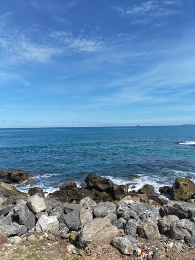 The image depicts a coastal scene in Naga Umbang, Indonesia. The foreground consists of a pile of large rocks, primarily gray and tan in color, extending to the shoreline. Small patches of green vegetation are visible among the rocks. The midground shows the ocean, with visible waves crashing against the rocks. The water transitions from turquoise near the shore to a deeper blue further out. In the distance, a ship is visible on the horizon, slightly left of center. An island is faintly discernible on the right side of the horizon. The sky occupies the upper portion of the image, featuring a bright blue color with scattered white, wispy clouds.