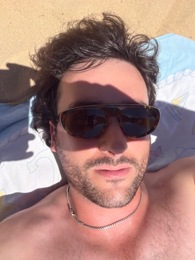 A man wearing sunglasses lies on his back. Part of his face is obscured by shadows and the sunglasses, which are reflected in the lenses. He has dark, wavy hair and a neatly trimmed beard. A silver chain adorns his neck. He is lying on a light blue towel with yellow and white sailboat patterns. The background appears to be sandy. The man's skin is tanned and there is visible body hair on his chest.