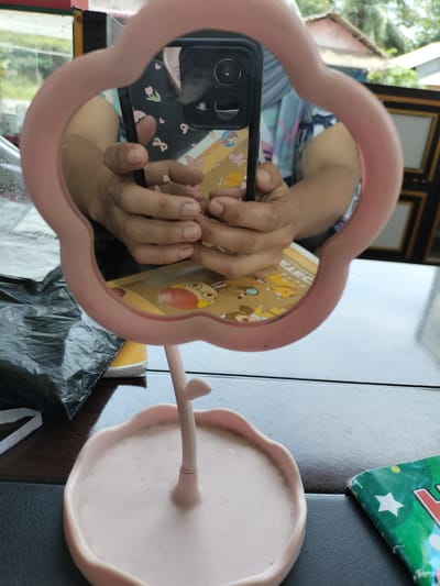 The image shows a pink flower-shaped mirror reflecting a person holding a smartphone. The person is wearing a patterned shirt with blue and white colors. The smartphone has a black casing with a triple camera system. In the background, the reflection shows some buildings and greenery, suggesting an outdoor or semi-outdoor setting. The mirror is placed on a table, and next to it, a black plastic bag and a green object with white stars are visible on the table. The overall scene appears to be in a domestic or commercial setting in Rantopanjang, Indonesia.