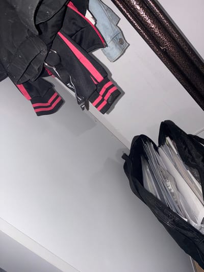 The image shows a cluttered closet interior. On the left, a black jacket with pink stripes is hanging. Attached to the jacket is a keychain with a metal ring and a black tag with white lettering that reads "FASHION". Next to the jacket, a light blue denim jacket is partially visible. On the right side of the image, a black open bag is filled with papers and documents. The closet appears to be located in an indoor residential setting in Rakitovo, Bulgaria. The lighting is dim, suggesting it may be nighttime or the closet is not well-lit. There are no people or animals visible in the image.